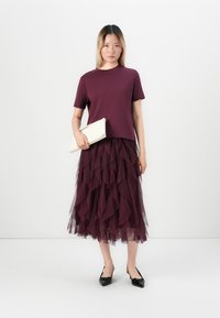 Woman wearing a short-sleeve purple top and layered purple tulle midi skirt, holding a white clutch and wearing black pointed-toe heels.