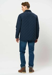 Homme portant une veste matelassée bleu foncé, un jean bleu et des bottes en cuir marron, debout de dos sur un fond clair uni.