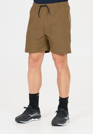 Beine mit braunen Shorts mit Kordelzug, schwarzen sportlichen Crew-Socken und schwarzen Sportschuhen, die auf weißem Hintergrund stehen.