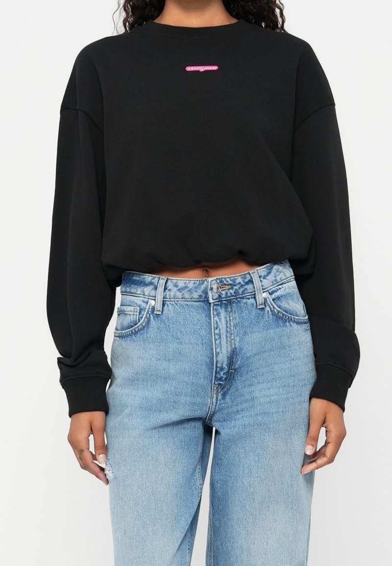 Femme portant un sweat-shirt court noir avec le logo "tommy jeans" et un jean denim bleu clair taille haute.