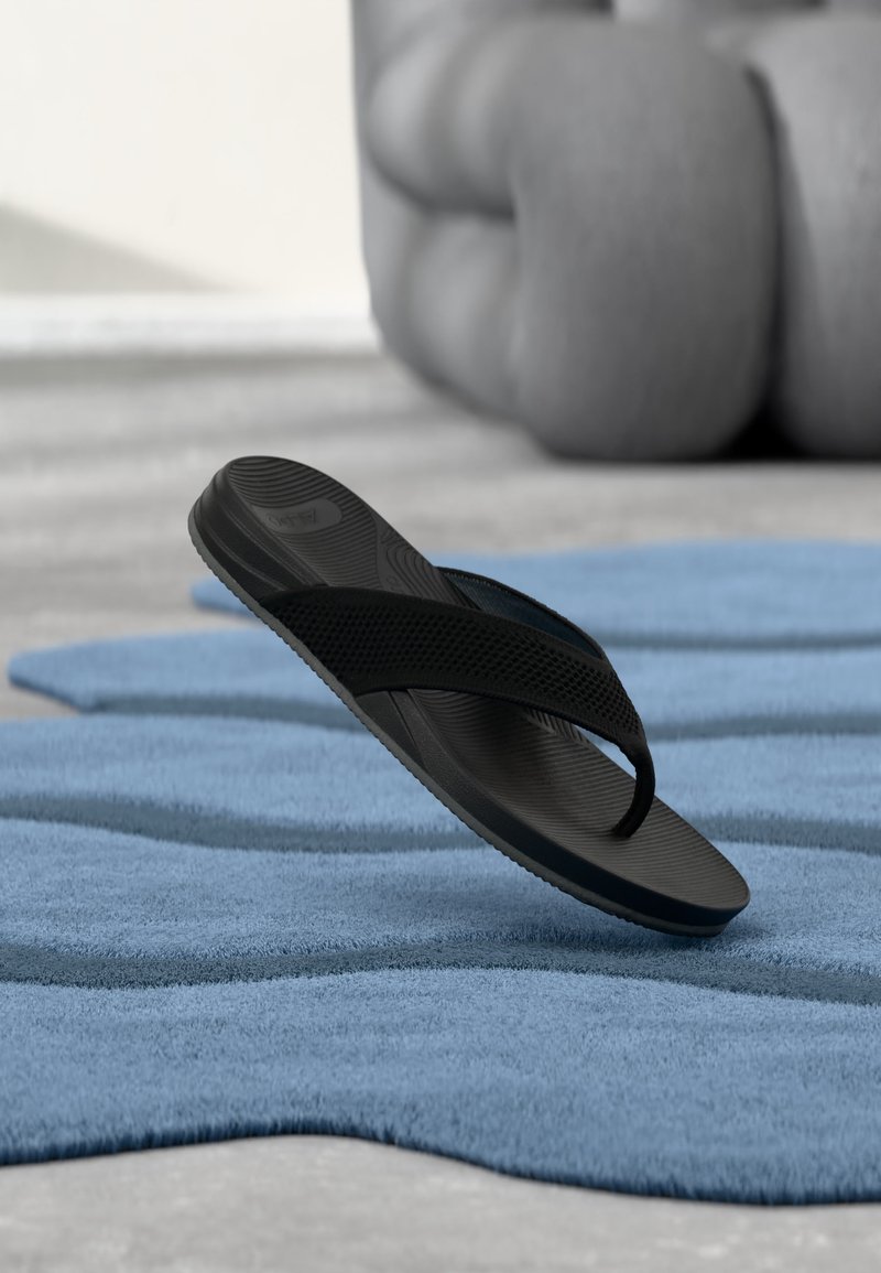 Fekete gumi papucs egy texturált kék szőnyegen, háttérben egy nagy, elmosódott szürke dísztárggyal.