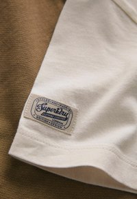 Tissu de couleur crème avec une étiquette rectangulaire cousue portant l'inscription « SuperDry Clothing Company London British Design » sur une surface texturée marron.