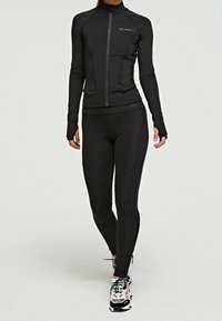 Chaqueta atlética negra con cuello alto y cremallera, combinada con leggins negros. Ambos presentan un tejido suave y elástico y un diseño ajustado.