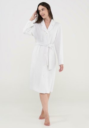 Mujer de pie descalza, vistiendo una bata blanca hasta la rodilla con botones y un cinturón atado, tocándose el cabello con una mano.