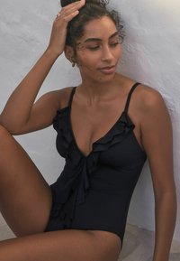 Black one-piece swimsuit with ruffled accents on the neckline, adjustable straps, and smooth fabric texture.