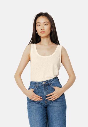 Jeune femme aux longs cheveux foncés portant un débardeur crème et un jean bleu taille haute, debout les mains dans les poches devant un fond blanc.