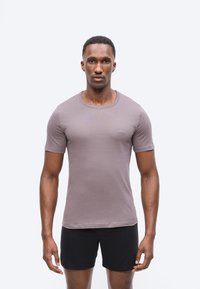 Αθλητικός άντρας που στέκεται αντιμετωπίζοντας μπροστά, φορώντας ένα εφαρμοστό πουκάμισο σε χρώμα μαβί και μαύρα σορτς, μπροστά σε ένα απλό φωτεινό φόντο.