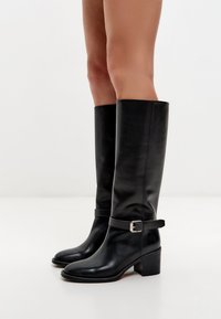 Cizme din piele neagră până la genunchi, cu vârf pătrat, toc bloc și o cataramă pe breteaua de la gleznă. Textură netedă și design minimalist.