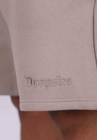 Beige shorts med glat tekstur, prydet med broderet "Drowsize" logo i en dekorativ font nær kanten. Klassisk snit med skjulte sømme.
