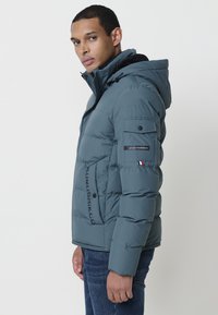 Modra puffer jakna s kapuco, teksturirana površina, zadrga spredaj in logotipni detajli na rokavu in strani. Obujena čez kavbojke.
