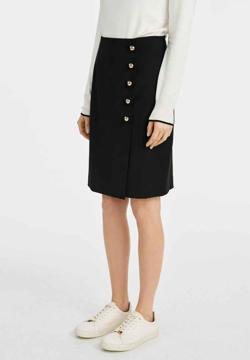 Czarna spódnica do kolan z przednim zapięciem na guziki, zawierająca pięć złotych guzików; gładki materiał o delikatnym połysku; noszona z białymi trampkami.