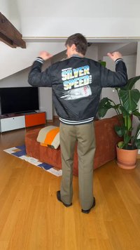 Άτομο που φοράει μαύρο μπουφάν με το κείμενο «Silver Speed» και γραφικό με κουτάκι στο πίσω μέρος, στέκεται σε ένα σύγχρονο σαλόνι με ξύλινο πάτωμα.