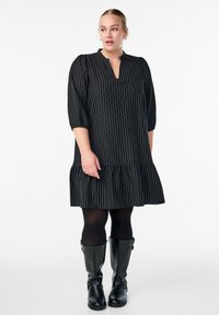 Nainen, jolla on päällä musta raidallinen mekko, jossa on 3/4-hihat, mustat legginsit ja mustat polvipitkät saappaat, seisoo yksinkertaista valkoista taustaa vasten.