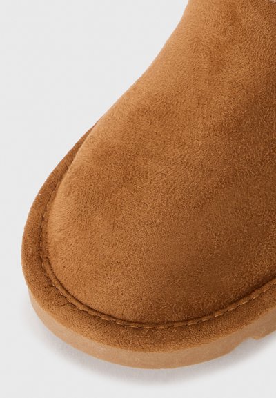Brun mockaläderstövel med rund tå, sydd kant och en platt gummisula. Mjuk textur, minimalistisk design utan ytterligare dekorationer.