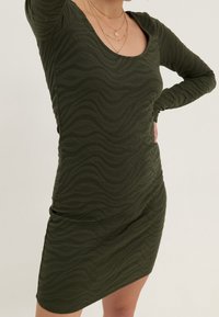 Robe longue à manches longues de couleur vert olive avec un motif texturé en forme de vague, encolure dégagée et silhouette ajustée, finissant à mi-cuisse. Colliers dorés en couches visibles.