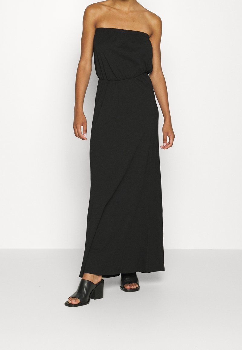 Femme portant une robe longue noire bustier avec une taille élastique et des mules noires à talons ouverts, debout devant un fond uni.