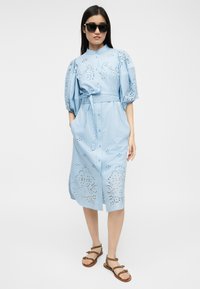 Γυναίκα που φοράει ανοιχτό μπλε midi φόρεμα με κουμπιά και σχέδια από δαντέλα με τρύπες, φουσκωτά μανίκια, ζώνη, καφέ σανδάλια και μαύρα γυαλιά ηλίου, όρθια.