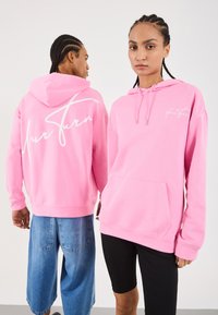 Zwei Personen mit geflochtenen Haaren tragen pinke Hoodies; eine steht frontal mit schwarzen Shorts, die andere zeigt die Rückseite des Hoodies mit blauen Denim-Shorts.
