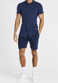 Tummansininen lyhythihainen polo-paita, jossa on napit ja logo rinnassa, yhdistettynä samansävyisiin shortseihin. Asusteena ovat valkoiset urheilusandaalit ja nilkkasukat.