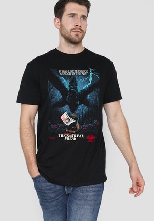 Man wearing black T-shirt with blue and red graphic of large shadowy creature, sailboat, and text referencing "Stranger Things" Netflix series.