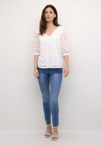 Hvid blonde top med trekvartærmer, V-udskæring, struktureret blomstermønster; parret med lyseblå skinny jeans og hvide strop sandaler.
