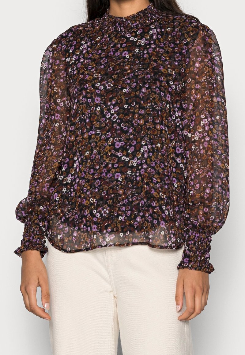 Blouse longue transparente à manches longues avec un petit motif floral violet, marron et blanc, col et poignets à volants, portée avec un pantalon blanc.