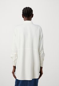 Camicia bianca a maniche lunghe con vestibilità rilassata, caratterizzata da un dettaglio della cucitura posteriore e polsini con bottoni. Tessuto morbido, design minimale.
