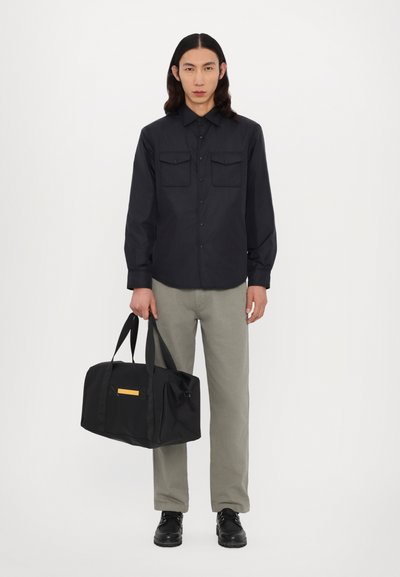 Black button-up shirt with two chest pockets, gray trousers, and a black duffel bag featuring a yellow accent. Smooth fabric, relaxed fit.