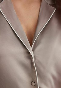 Sidenblus i taupe-färg, med kontrasterande vit kant längs kragen och knappstängning fram. Slät textur.