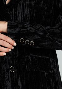 Blazer in velluto nero con tessuto testurizzato, revers a punta e dettagli con bottoni argentati sui polsini e sul davanti. Dettaglio delle tasche visibile.
