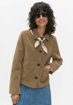 Kvinde med krøllet hår iført sorte solbriller, en brun jakke med knapper, cremefarvet mønstret halstørklæde og blå denim nederdel, smilende.