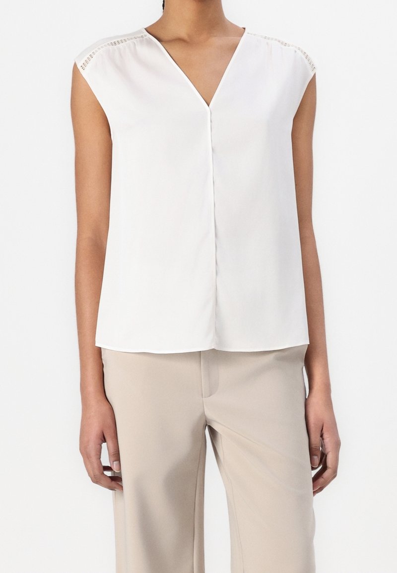 Blouse blanche sans manches avec un col en V, présentant de subtils accents aux épaules et une texture lisse, associée à un pantalon beige clair.