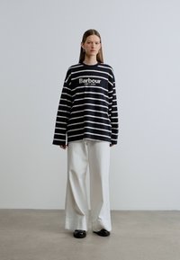 Zwart-wit gestreept shirt met lange mouwen en een logo op de borst. Gecombineerd met witte broek met wijde pijpen en zwarte schoenen. Minimalistische stijl.