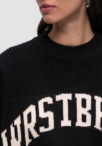 Schwarzer Strickpullover mit einer dicken Rippenstruktur, der weiße Blockbuchstaben aufweist. Der Kragen ist rund und eng anliegend.