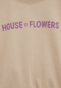 Bézs pulóver bordázott nyakkivágással, rajta nagy, texturált lila betűkkel a "HOUSE of FLOWERS" felirat. Puha szövet textúra.