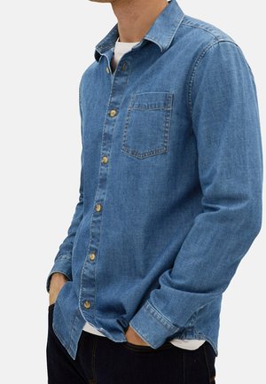 Chemise en denim bleu avec boutonnage à l'avant, col rabattu, poche poitrine unique et manches longues. Présente des boutons dorés et des coutures détaillées.