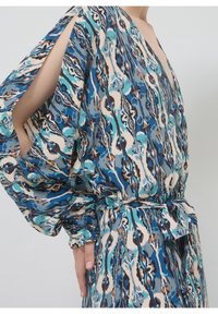 Robe fluide avec motif abstrait bleu, beige et marron, dotée d’un décolleté en V profond, d’une taille nouée et de manches fendues laissant les bras nus.