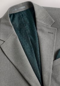 Gros plan sur un blazer gris texturé avec une doublure intérieure à motif vert foncé et un mouchoir de poche assorti visible dans la poche poitrine.