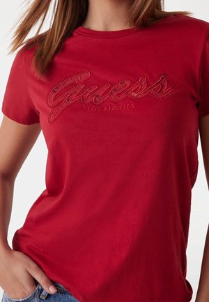 Femme portant un t-shirt rouge avec l'inscription "Guess Los Angeles" brodée sur la poitrine, associé à un jean en denim bleu.