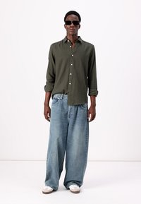 Mörkgrön linneskjorta med knappar, löst sittande ljusblå denimjeans, svarta solglasögon och vita sneakers. Enkelt design, avslappnad stil.