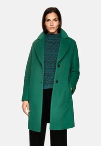 Grüner Wollmantel mit großen Revers und zwei Knöpfen. Darunter ein strukturiertes, mehrfarbig gestreiftes Sweatshirt in Blau, Braun und Schwarz.