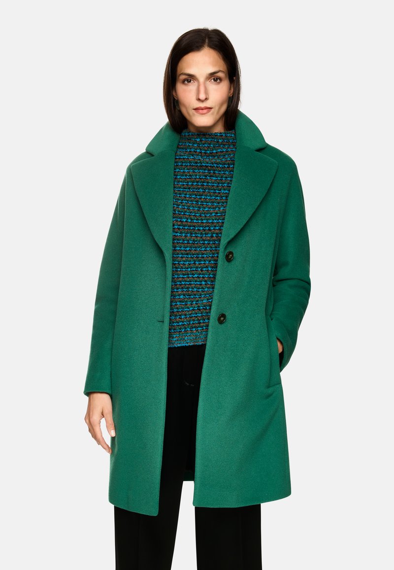 Grüner Wollmantel mit großen Revers und zwei Knöpfen. Darunter ein strukturiertes, mehrfarbig gestreiftes Sweatshirt in Blau, Braun und Schwarz.