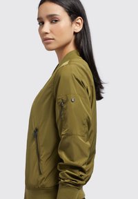 Olive rohelise värvi bomber-jakk, millel on lukuga küljetasku, soonikkoes mansetid ja õlavarre detailid. Valmistatud siledast, kergest kangast.