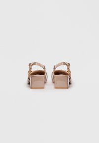 Pari beige ki patent leather block heel sandals jo ankle straps ke saath hain, seedhe peeche se dikhai de rahe hain ek saaf safed background par.