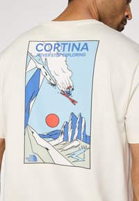 Μπλουζάκι από βαμβάκι σε κρεμ χρώμα με πολύχρωμο γραφικό σχέδιο που απεικονίζει βουνά, έναν σκιέρ και έναν ήλιο. Το κείμενο γράφει "CORTINA NEVER STOP EXPLORING."