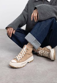 Zapatillas altas de lona beige con cordones blancos y suela de goma. Presentan costuras en contraste y un logotipo en la lengüeta, sobre un fondo de denim.