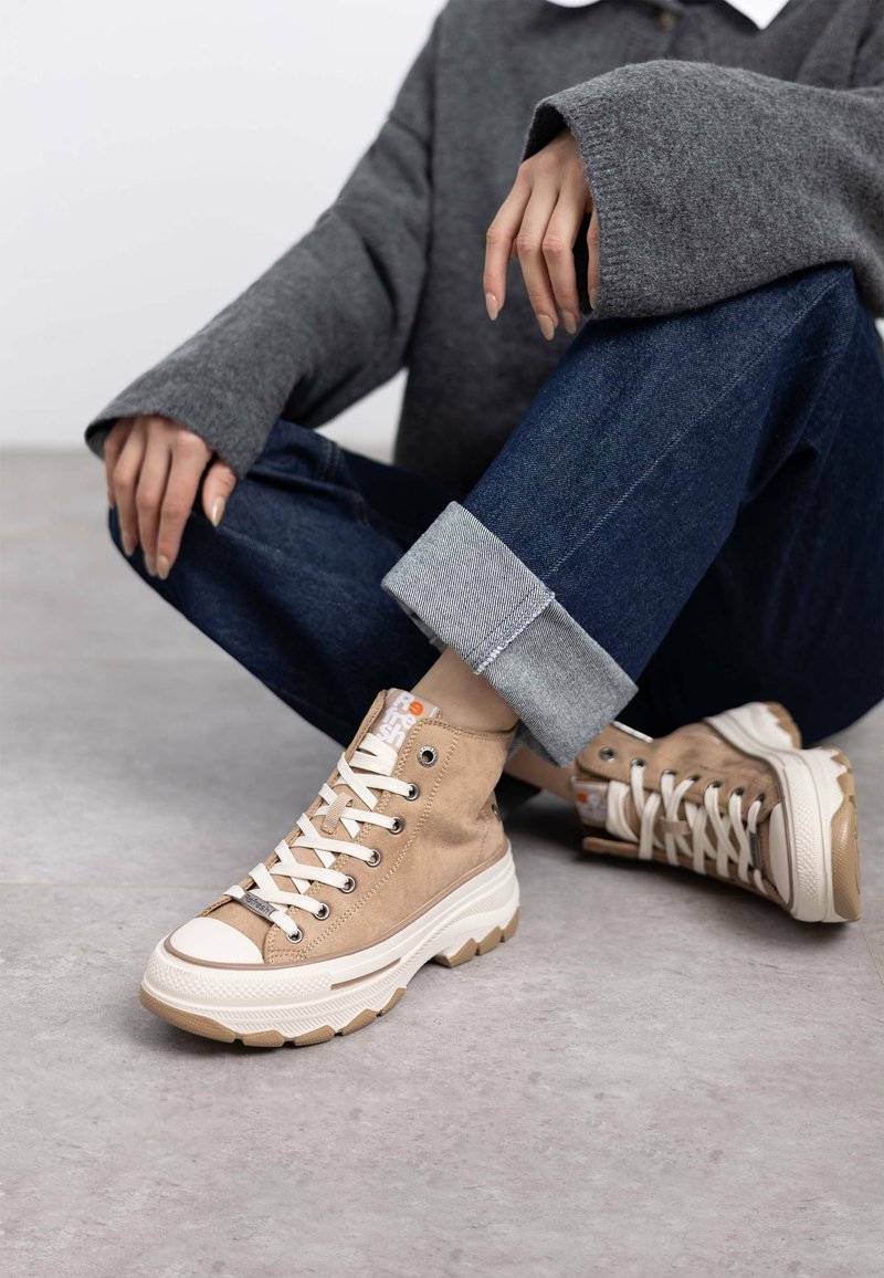 Zapatillas altas de lona beige con cordones blancos y suela de goma. Presentan costuras en contraste y un logotipo en la lengüeta, sobre un fondo de denim.