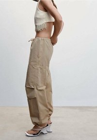 Femme portant un pantalon cargo beige, un top court en perles et des sandales à talons blancs à lanières, se tenant de profil avec les mains derrière le dos.