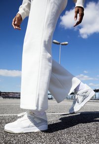 Chaussures de sport blanches avec des accents gris, surfaces texturées et lacets. Associées à un pantalon blanc ample, sur un fond de ciel bleu.