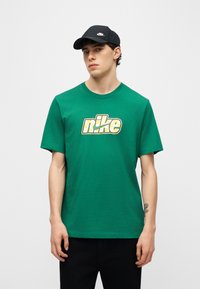 Jeune homme portant un t-shirt Nike vert avec un logo jaune et une casquette Nike noire, debout devant un fond clair uni.